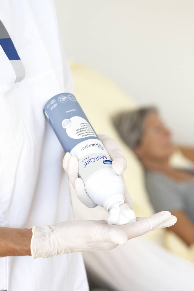Bildbeschreibung: Auf dem Bild ist der MoliCare Skin Reinigungsschaum zu sehen. Der Produktbehälter hat eine schlanke, weiße Flasche mit einem blauen Pumpkopf. Der Schaum wird aus der Flasche gepumpt und hat eine leichte, luftige Konsistenz. Im Hintergrund ist eine stilvolle, helle Umgebung zu erkennen, die einen hygienischen und modernen Eindruck vermittelt. Neben der Flasche sind einige handliche Tücher oder einen Schwamm zu sehen, die zur Anwendung des Schaums verwendet werden können.