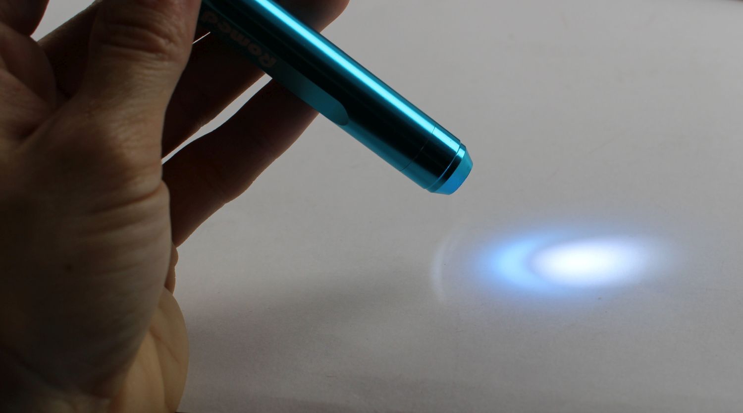 Hand hält eingeschaltete Romed Diagnostikleuchte, heller weißblauer Lichtstrahl auf grauem Untergrund